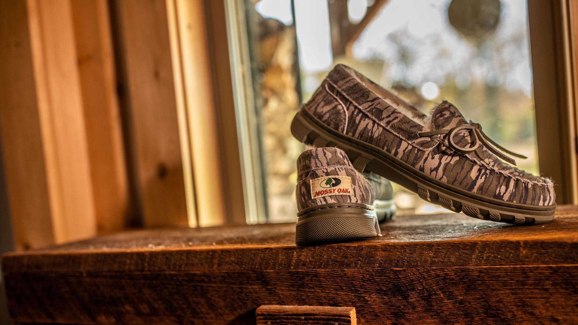 BoneZZZ Men's Flex Slippers in Mossy Oak Bottomland pattern, sitting on a wooden trunk.