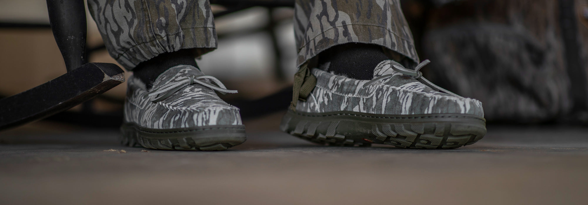 Close-up of a man wearing his hunting camouflage pants and his BoneZZZ Men's Flex Slippers, in Mossy Oak Bottomland pattern, on his front porch.