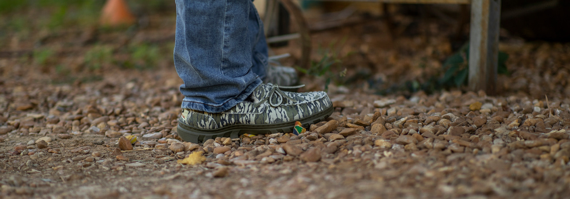 Close-up of a man wearing jeans and his BoneZZZ Men's Flex Slippers, in Mossy Oak Bottomland pattern, outside in the yard on gravel.