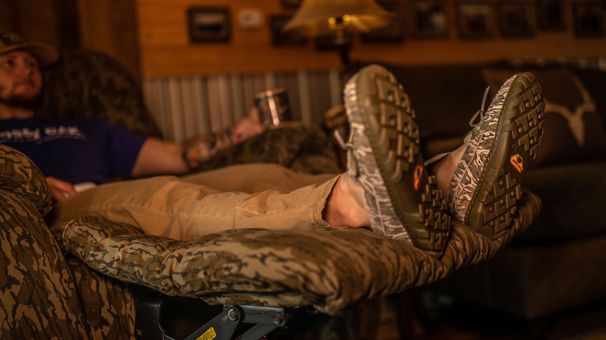 Close-up of a man relaxing in a recliner with his feet up while wearing his BoneZZZ Men's Flex Slippers, in Mossy Oak Bottomland pattern.
