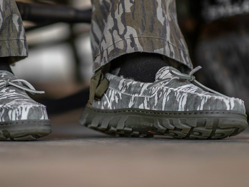 Close-up of a man wearing his hunting camouflage pants and his BoneZZZ Men's Flex Slippers, in Mossy Oak Bottomland pattern, on his front porch.