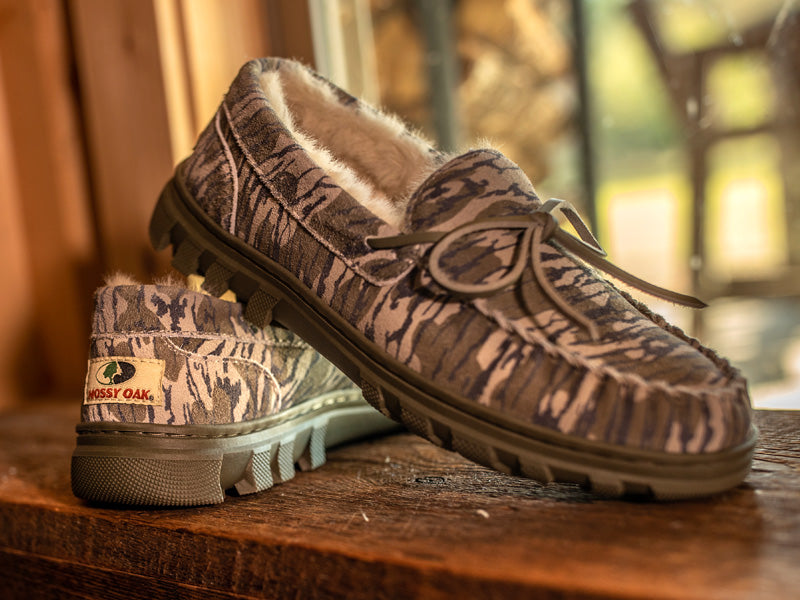 BoneZZZ Men's Flex Slippers in Mossy Oak Bottomland pattern, sitting on a wooden trunk.